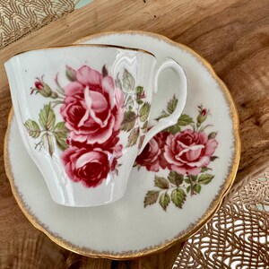 Pu&ograve; includere: Una tazza da t&egrave; e un piattino bianchi con un motivo floreale di rose rosa e foglie verdi. La tazza e il piattino hanno un bordo dorato.