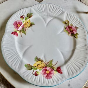 May include: White ceramic plate with a scalloped edge and floral design. The plate features pink, yellow, and red flowers with green leaves. The plate is on a heart-shaped tray with gold handles.