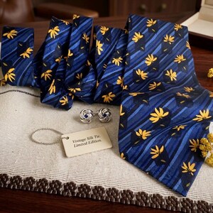 May include: A navy blue silk tie with a yellow floral pattern and diagonal stripes. The tie is accompanied by silver cufflinks and a tag that reads "Vintage Silk Tie. Limited Edition."