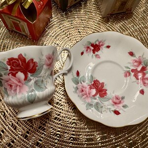 May include: A white teacup and saucer set with a floral design featuring pink and red roses. The set has a gold trim around the edges.