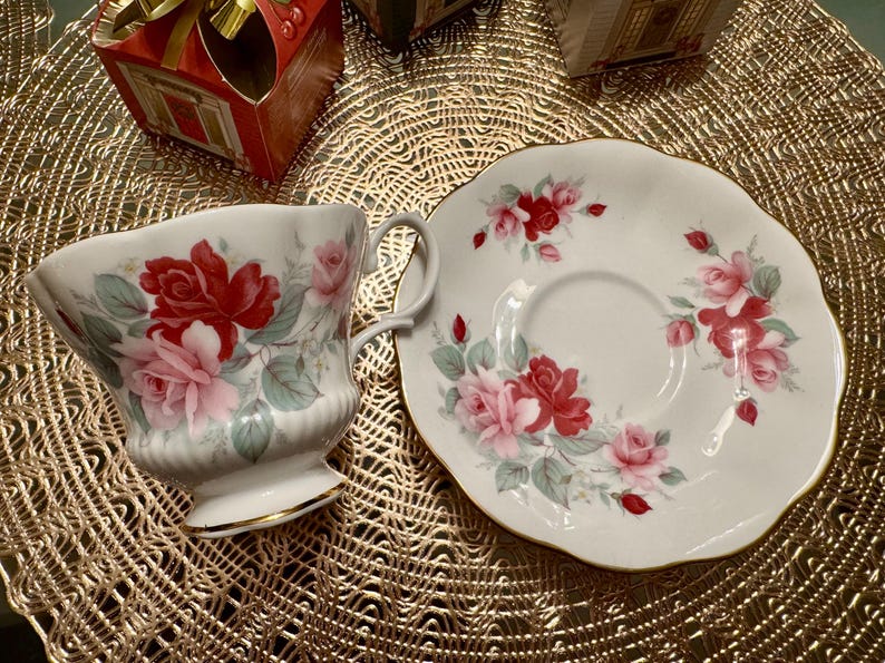 May include: A white teacup and saucer set with a floral design featuring pink and red roses. The set has a gold trim around the edges.
