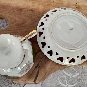 Puede incluir: Juego de taza y platillo blancos con ribete dorado. El platillo tiene recortes en forma de coraz&oacute;n en el borde. La taza y el platillo est&aacute;n sobre una superficie de madera, lo que sugiere un estilo vintage o antiguo.