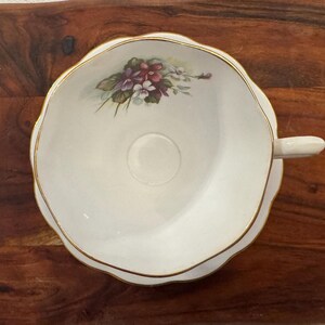 May include: A white teacup and saucer with a gold rim and a floral design featuring purple and pink flowers. The saucer has a scalloped edge.
