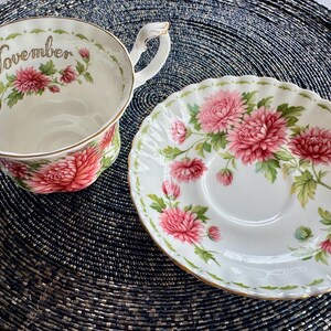 May include: A white teacup and saucer set with a floral design. The teacup has the word "November" printed on it. The set features pink flowers and green leaves with gold trim.