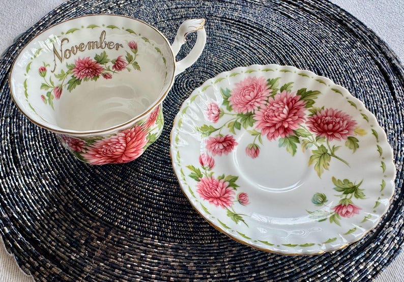 May include: A white teacup and saucer set with a floral design. The teacup has the word "November" printed on it. The set features pink flowers and green leaves with gold trim.