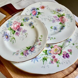 May include: Three white porcelain plates with a floral pattern. The plates have pink, purple, and blue flowers with gold edging. They are displayed on a wooden serving platter.