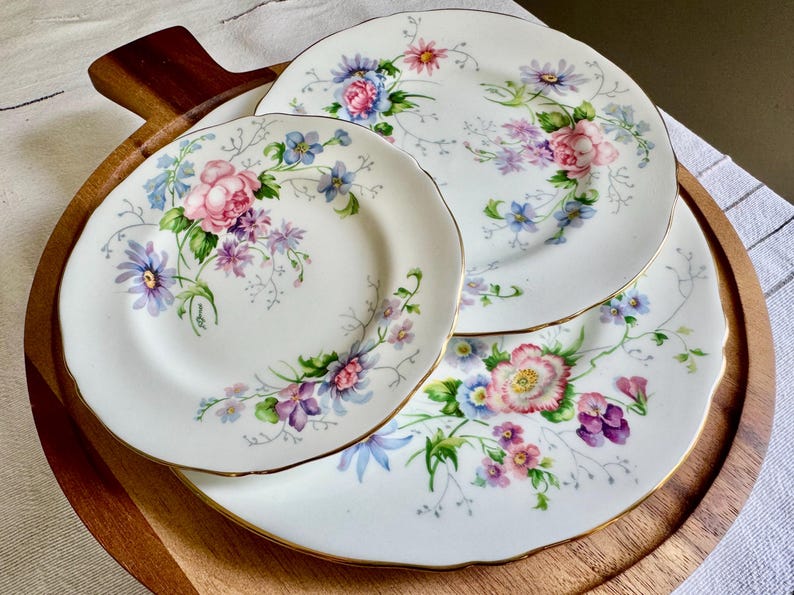 May include: Three white porcelain plates with a floral pattern. The plates have pink, purple, and blue flowers with gold edging. They are displayed on a wooden serving platter.