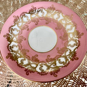 May include: A pink and white teacup saucer with gold filigree detailing. The saucer has a central white circle for the cup and a decorative gold and white pattern around the edge. The rim is also trimmed in gold.