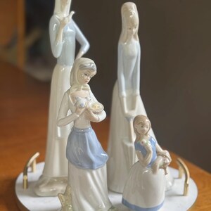 K&ouml;nnte beinhalten: Eine Sammlung von Porzellanfiguren, die Frauen und ein Kind darstellen. Die Figuren tragen lange, flie&szlig;ende Kleider mit pastellblauen und wei&szlig;en Akzenten. Die Figuren sind auf einem herzf&ouml;rmigen Marmortablett mit goldenem Rand angeordnet.