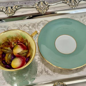 May include: A vintage teacup and saucer set. The teacup features a hand-painted design of peaches and grapes. The saucer is a solid light teal color with a gold rim. The set is displayed on a silver tray.