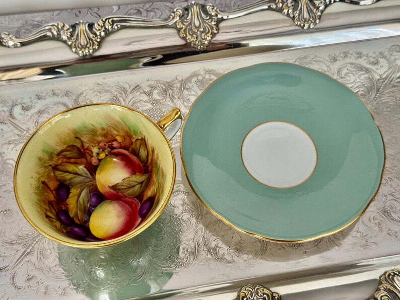 May include: A vintage teacup and saucer set. The teacup features a hand-painted design of peaches and grapes. The saucer is a solid light teal color with a gold rim. The set is displayed on a silver tray.