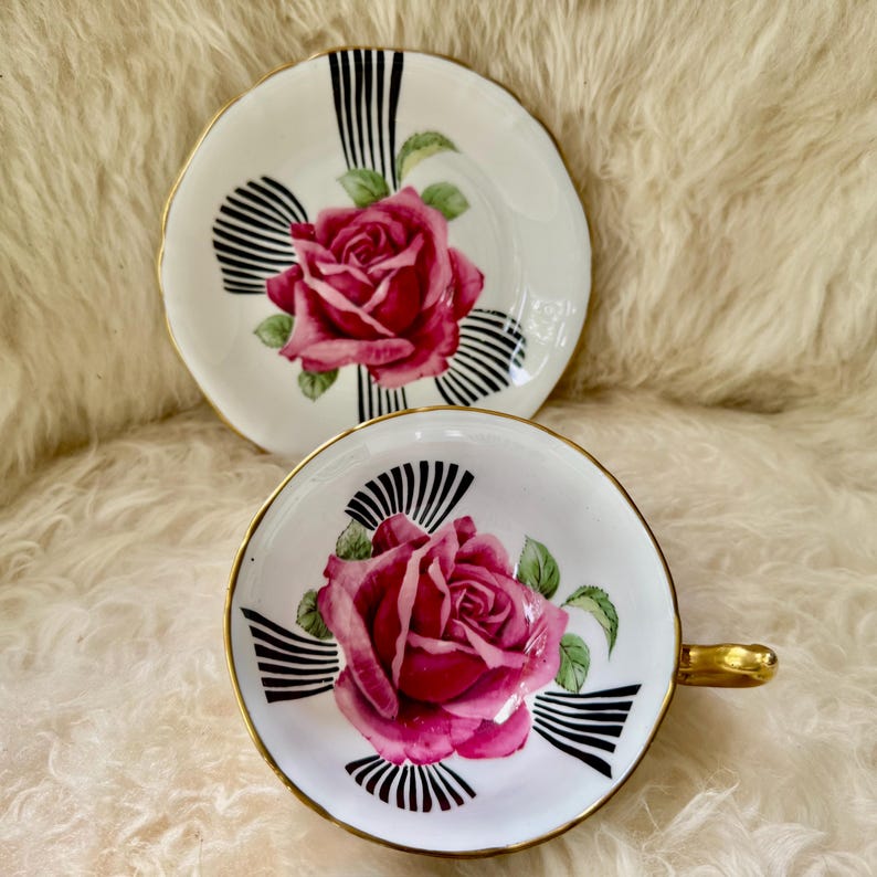 May include: A white teacup and saucer set with a pink rose design. The cup has a gold handle and trim, and the saucer has a matching gold rim. Black and white striped accents surround the rose, creating a vintage aesthetic.