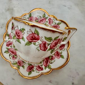 Puede incluir: Un juego de taza y platillo de t&eacute; vintage. La taza y el platillo de porcelana blanca est&aacute;n adornados con un dise&ntilde;o floral de flores rosas y hojas verdes. Los bordes de la taza y el platillo est&aacute;n acentuados con detalles dorados.