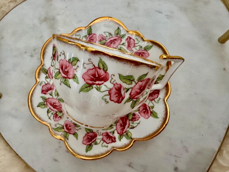 Puede incluir: Un juego de taza y platillo de t&eacute; vintage. La taza y el platillo de porcelana blanca est&aacute;n adornados con un dise&ntilde;o floral de flores rosas y hojas verdes. Los bordes de la taza y el platillo est&aacute;n acentuados con detalles dorados.