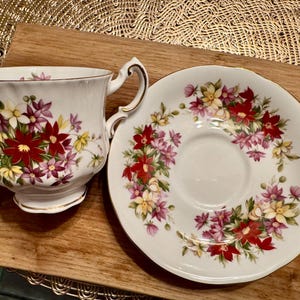 May include: A white teacup and saucer set with a floral pattern featuring red, pink, and yellow flowers. The set is decorated with a gold trim.