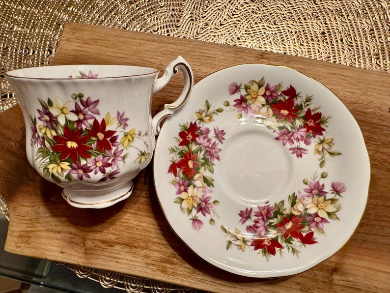 May include: A white teacup and saucer set with a floral pattern featuring red, pink, and yellow flowers. The set is decorated with a gold trim.