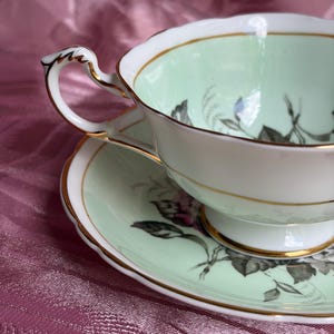 May include: A porcelain teacup and saucer set featuring a pale mint green interior and rim. The set is decorated with black and white floral illustrations and finished with a delicate gold trim along the scalloped edges and the ornate handle.