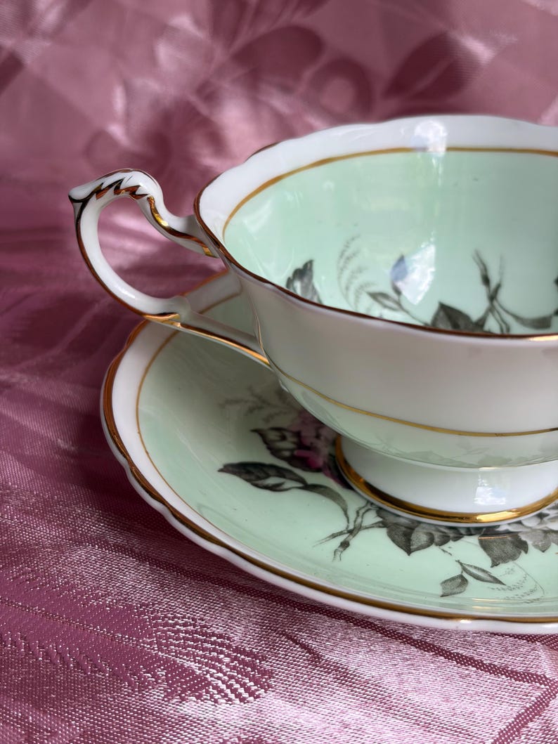 May include: A porcelain teacup and saucer set featuring a pale mint green interior and rim. The set is decorated with black and white floral illustrations and finished with a delicate gold trim along the scalloped edges and the ornate handle.