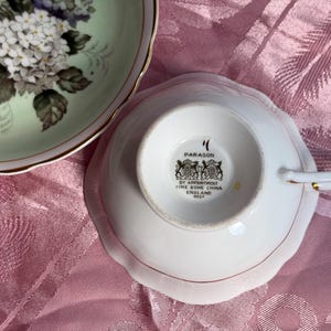 May include: A white fine bone china teacup by Paragon with a gold-trimmed handle, shown upside down to reveal the maker's mark and crest. Beside it sits a matching saucer featuring a pale green center decorated with a cluster of white and purple flowers.