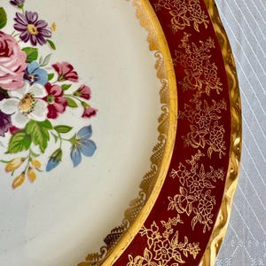 May include: A vintage decorative plate with a floral design. The plate features a white center with a bouquet of pink, purple, and blue flowers. The rim is a deep red with gold floral patterns and a gold scalloped edge.