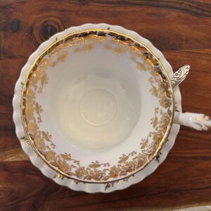 Puede incluir: Vista superior de una taza y platillo blancos con detalles florales dorados. Una peque&ntilde;a cuchara plateada descansa sobre el platillo. El conjunto est&aacute; sobre una superficie de madera oscura.