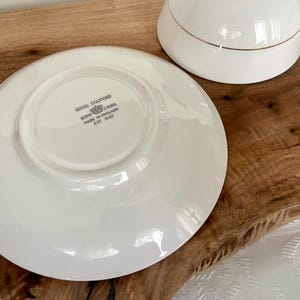 Pode incluir: Um conjunto de ch&aacute; branco com detalhes dourados. O pires est&aacute; na parte inferior, com a ch&aacute;vena de ch&aacute; &agrave; direita. O pires tem o texto "Royal Stafford Bone China Made in England Est 1845".