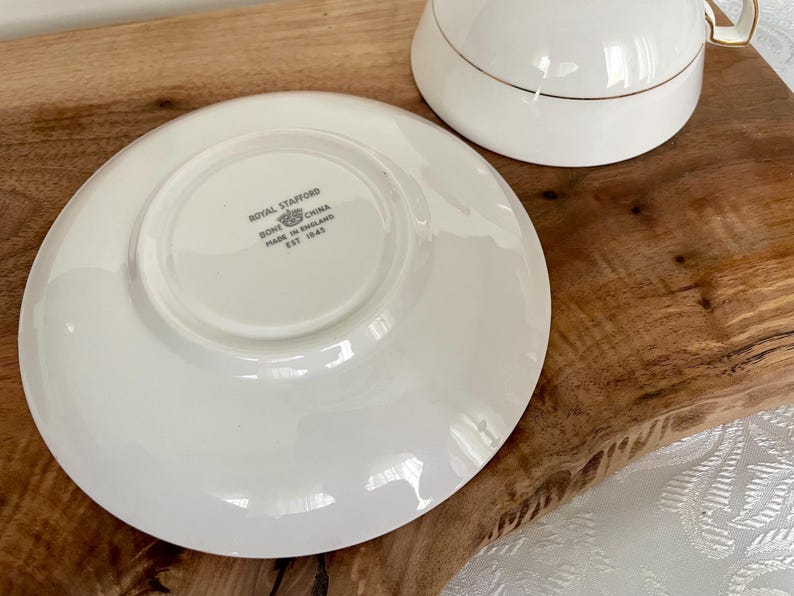 Pode incluir: Um conjunto de ch&aacute; branco com detalhes dourados. O pires est&aacute; na parte inferior, com a ch&aacute;vena de ch&aacute; &agrave; direita. O pires tem o texto "Royal Stafford Bone China Made in England Est 1845".