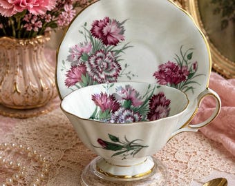 May include: A white porcelain teacup and matching saucer featuring a delicate floral pattern of deep pink carnations with green stems and leaves. The set has a scalloped edge, a gold-trimmed handle, and a gold-rimmed base on the teacup.
