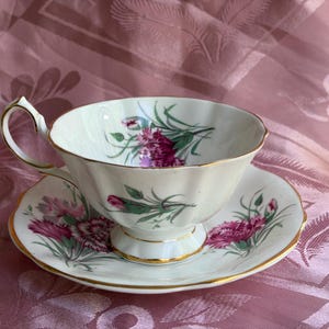 May include: A white porcelain teacup and saucer set featuring a fluted edge and gold trim. The set is decorated with a floral pattern of pink and magenta carnations with green stems and leaves, painted on both the interior and exterior surfaces.
