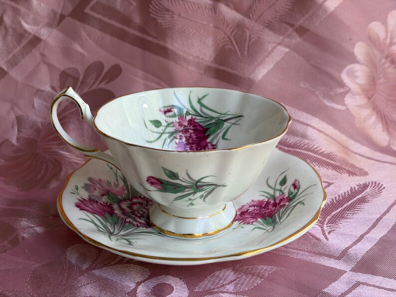 May include: A white porcelain teacup and saucer set featuring a fluted edge and gold trim. The set is decorated with a floral pattern of pink and magenta carnations with green stems and leaves, painted on both the interior and exterior surfaces.