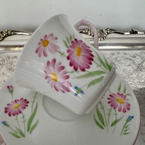 May include: White teacup and saucer set with a floral daisy design. The daisies have pink petals and yellow centers, with green leaves and blue accents. The cup has a pink handle and rim.