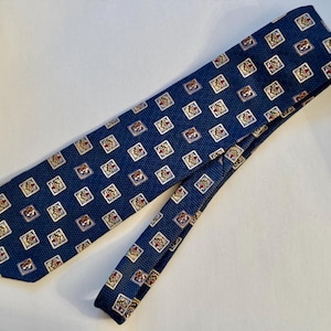 May include: A navy blue necktie featuring a repeating pattern of small white squares, each containing a centered gold crown icon with red accents. The fabric has a fine, textured weave throughout the entire length of the necktie.