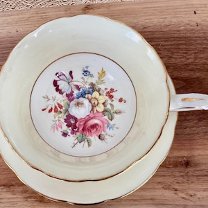May include: A vintage teacup and saucer set with a floral design. The cup and saucer are both white with a delicate gold trim. The floral design features a bouquet of pink, yellow, and purple flowers.