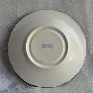 May include: The underside of a white bone china saucer with a scalloped edge. The center features a blue stamp that reads Royal Stafford, Bone China, Made in England. The surface is smooth and circular with a raised foot rim in the center.
