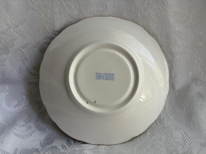 May include: The underside of a white bone china saucer with a scalloped edge. The center features a blue stamp that reads Royal Stafford, Bone China, Made in England. The surface is smooth and circular with a raised foot rim in the center.
