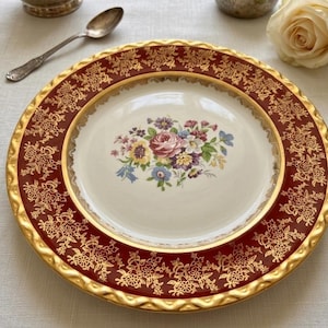 May include: A decorative plate with a maroon border featuring a gold floral pattern. The centre of the plate is cream-coloured with a floral bouquet design. The plate has a gold rim and is likely vintage or antique.