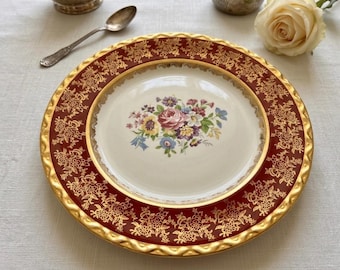 May include: A decorative plate with a maroon border featuring a gold floral pattern. The center of the plate is cream-colored with a floral bouquet design. The plate has a gold rim and is likely vintage or antique.