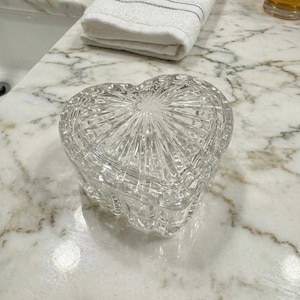 May include: A clear glass trinket box shaped like a heart, featuring a faceted, sunburst-like pattern radiating from the center of the lid. The container has vertical ribbed sides and a smooth, transparent finish throughout its entire structure.