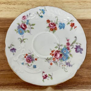 May include: White porcelain saucer with a scalloped edge and gold trim. The saucer is decorated with a floral pattern featuring pink, red, blue, and purple flowers. The design is centered around a recessed area for a teacup.