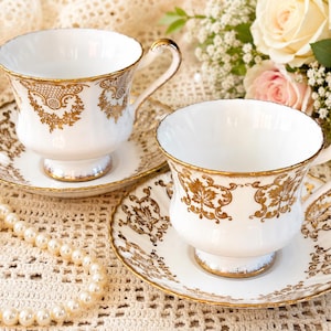 May include: Two white porcelain teacups and matching saucers featuring intricate gold filigree patterns along the rims and bodies. Each cup has a pedestal base and a curved handle, with gold trim detailing on the edges of the saucers and cups.