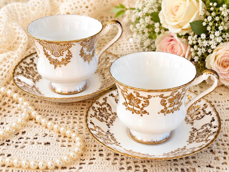 May include: Two white porcelain teacups and matching saucers featuring intricate gold filigree patterns along the rims and bodies. Each cup has a pedestal base and a curved handle, with gold trim detailing on the edges of the saucers and cups.