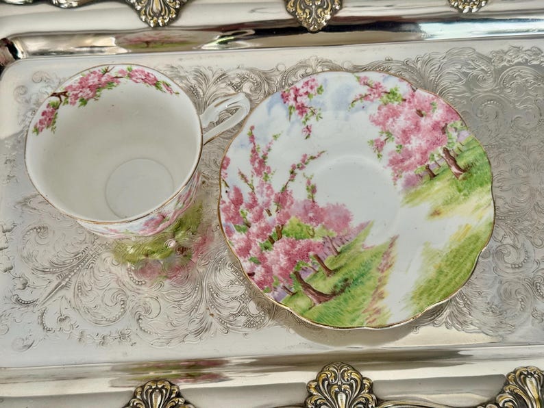 May include: A white teacup and saucer set with a floral design featuring pink blossoms and green foliage. The set is placed on a silver tray with ornate detailing, creating an elegant presentation.