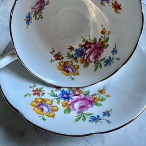 Puede incluir: Un juego de taza y platillo blancos con un dise&ntilde;o floral. La taza y el platillo tienen un borde festoneado y ribete dorado. El patr&oacute;n floral incluye rosas rosas, flores azules, amarillas y rojas.