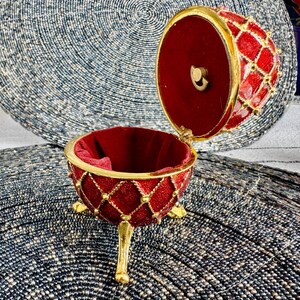 May include: A red and gold jeweled egg-shaped trinket box with a hinged lid and three gold legs. The box is open and the inside is lined with red velvet.