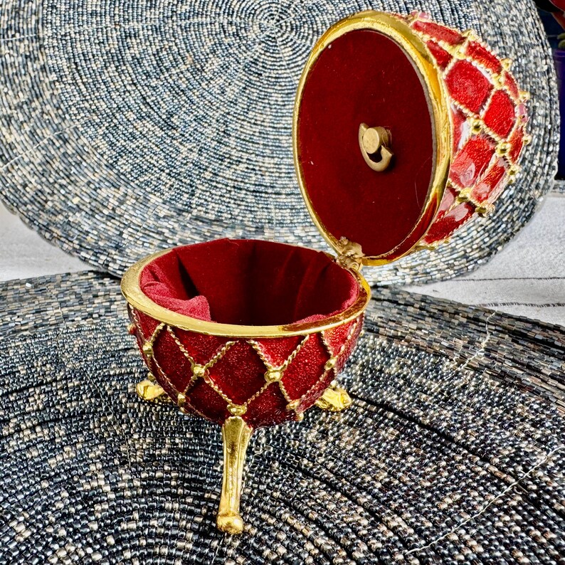 May include: A red and gold jeweled egg-shaped trinket box with a hinged lid and three gold legs. The box is open and the inside is lined with red velvet.