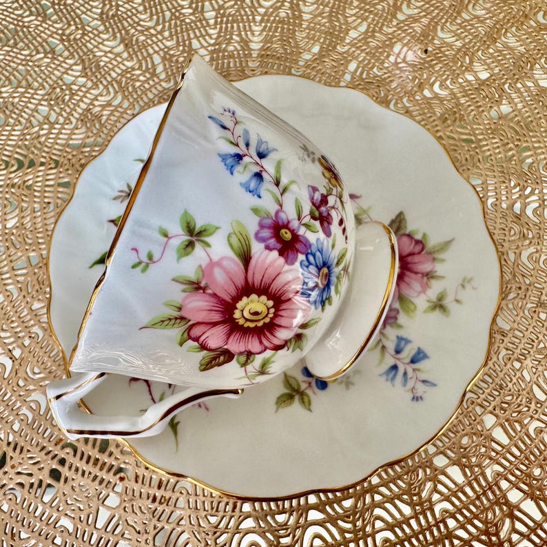 May include: A white teacup and saucer with a floral pattern in shades of pink, purple, and blue. The cup and saucer have a gold trim.