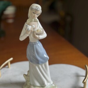 K&ouml;nnte beinhalten: Porzellanfigur einer Frau in wei&szlig;er Bluse und blauem Rock, die einen Vogel und einen Korb h&auml;lt. Die Figur hat einen ruhigen Ausdruck und steht auf einer marmor&auml;hnlichen Oberfl&auml;che.