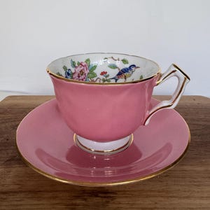 May include: A pink teacup and saucer set. The teacup features a floral and bird design inside the cup, with gold trim. The saucer and cup are a solid pink color with gold trim. The handle is white with gold accents.
