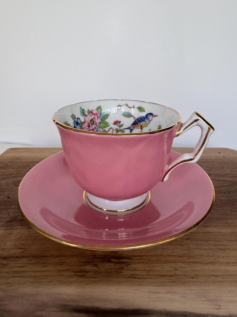 May include: A pink teacup and saucer set. The teacup features a floral and bird design inside the cup, with gold trim. The saucer and cup are a solid pink color with gold trim. The handle is white with gold accents.