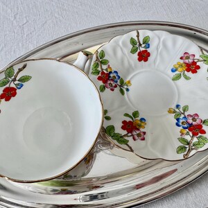 K&ouml;nnte beinhalten: Ein wei&szlig;es Tassen- und Untertassenset mit einem Blumenmuster in Rot, Gelb und Blau, das auf einem silbernen ovalen Tablett liegt. Die Tasse hat einen Goldrand und ein passendes Blumenmuster.